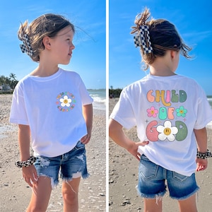 May include: White t-shirt with a colorful graphic of a daisy and the text "Child of God" on the front. The back of the shirt has a similar graphic with the text "Child of God" in a rainbow color scheme.