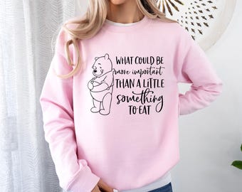 Könnte beinhalten: Rosafarbener Pullover mit einem schwarzen Aufdruck von Winnie Puuh, der ein Honigglas hält. Der Text lautet "What could be more important than a little something to eat."