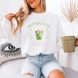 May include: White sweatshirt featuring a Matcha Latte graphic. The design includes a green matcha drink in a clear cup with a pink bow, surrounded by green leaves, and the text "Matcha Latte Social Club".