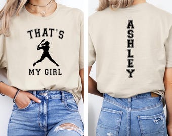 May include: A beige t-shirt with the text "That's My Girl" and a silhouette of a baseball player on the front. The back of the shirt has the name "Ashley" printed vertically down the back.