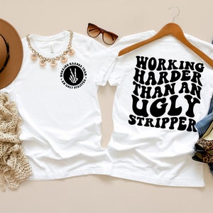 Puede incluir: Camiseta blanca con el texto "Working Harder Than An Ugly Stripper" en negro, fuente retro. La camiseta est&aacute; en una percha con un par de jeans y una planta de algod&oacute;n en el fondo.
