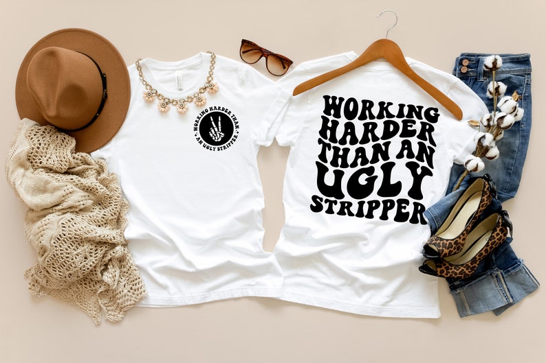 Puede incluir: Camiseta blanca con el texto "Working Harder Than An Ugly Stripper" en negro, fuente retro. La camiseta est&aacute; en una percha con un par de jeans y una planta de algod&oacute;n en el fondo.