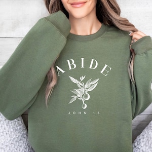 May include: A green sweatshirt with the word "ABIDE" and a small floral design printed on the front. The text "JOHN 15" is printed below the design.