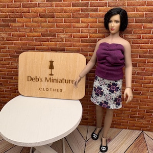 May include: A miniature outfit consisting of a purple tube top and a floral print skirt. The outfit is displayed on a doll. A wooden sign with the text "Deb's Miniature CLOTHES" is also visible.