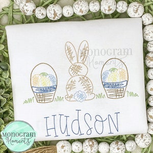 May include: Embroidered white t-shirt featuring a brown bunny rabbit, two baskets of colourful eggs, and the name "HUDSON" in blue thread.