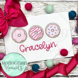 Könnte beinhalten: Weißes T-Shirt mit drei gestickten rosafarbenen und braunen Donuts. Der Name "Gracelyn" ist in rosa unter den Donuts gestickt.