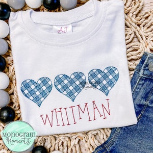 May include: White t-shirt with three blue and white checkered hearts and the name "WHITMAN" embroidered in red.