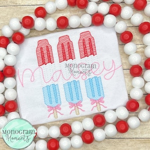 May include: A white t-shirt with red, white, and blue popsicle embroidery. The text "Mommy" is embroidered in pink above the popsicles. The t-shirt is surrounded by a string of red and white beads.