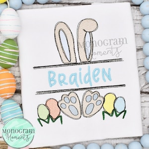 May include: A white embroidered shirt with a bunny design. The bunny has brown ears, a blue nose, and brown paws. The shirt also has a blue banner with the name "Braiden" embroidered on it. The bunny is surrounded by colorful Easter eggs.