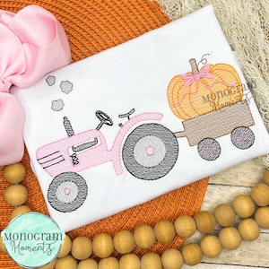 May include: A pink tractor pulling a wagon with a pumpkin on top. The pumpkin has a pink bow on it. The tractor and wagon are embroidered on a white shirt.