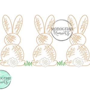 May include: Three brown bunny rabbits with a stitched design, sitting on a green grass background. The text "Monogram Moments" is in a circle above the bunnies.