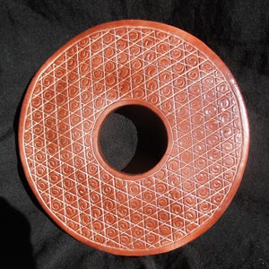 May include: A circular, reddish-brown object with a central hole and an intricate, repeating pattern of geometric shapes and spirals carved into its surface. The object is set against a dark background, highlighting the detailed design.