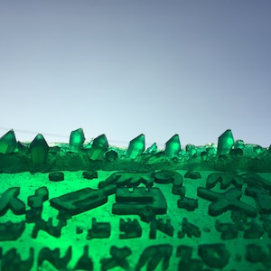 May include: A close-up of a green, crystal-like surface with a textured, rough appearance. The surface is covered in small, green crystals that create a jagged, uneven edge.