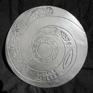 May include: A silver-colored, circular decorative plate with an embossed spiral design. The design includes a lizard, sun, and other stylized figures. The plate also features an inscription around the bottom edge.