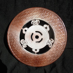 May include: A circular, decorative object with a brown outer ring and a white, central design. The brown ring features a repeating swirl pattern. The white center has a circular design with a central hole and four decorative elements.