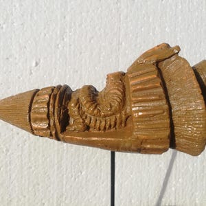 Könnte beinhalten: Eine detaillierte, braune, geschnitzte Holzskulptur eines Projektils, möglicherweise einer Kugel oder einer stilisierten Waffe. Die Skulptur hat eine spitze Spitze und komplizierte, strukturierte Details entlang ihres Körpers, mit einem zylindrischen Sockel und einem abgerundeten Ende.