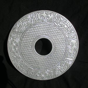 May include: A silver-colored, circular decorative object with an embossed design. The outer edge features intricate scrollwork and patterns, surrounding a central area with a diamond grid texture and a large central hole. The object is likely made of metal.