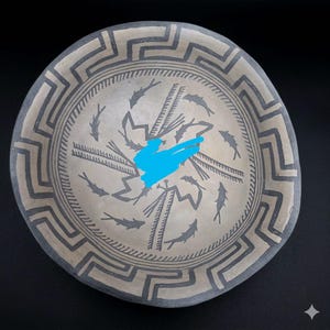May include: A round, shallow ceramic bowl with a light gray surface featuring dark gray painted details. The rim displays a continuous geometric meander pattern, while the interior shows a circular arrangement of fish motifs and jagged lines.