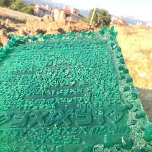 May include: A green resin plaque with a raised design of a script. The plaque is surrounded by a border of green crystals.