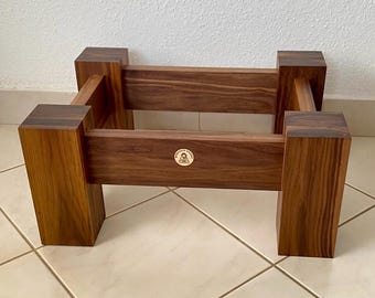 May include: A rectangular wooden stand featuring four thick corner posts and horizontal support beams. The wood displays a rich brown grain pattern. A small circular logo with a graphic of a seated figure and the text BUDDHOP MONK is centered on the front beam.