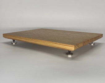 May include: A rectangular wooden platform with a natural brown wood grain finish. The platform is elevated by four small, conical black feet that are attached to circular silver-toned metal bases, providing a stable and raised surface.