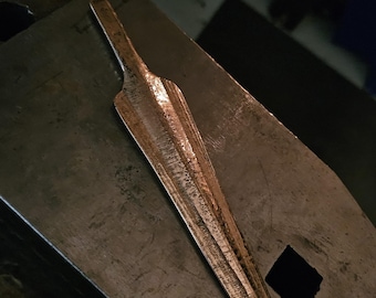 May include: A hand-forged metal spearhead with a pointed tip and a narrow tang. The surface of the metal displays a textured, dark, and weathered finish with visible hammer marks along the central ridge and edges of the blade.