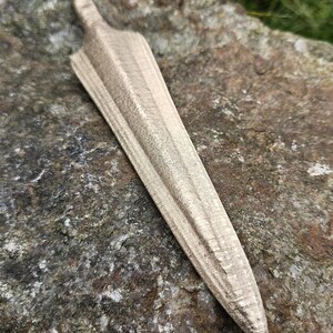 Puede incluir: Una daga de madera con una superficie texturizada, apoyada sobre una roca. La daga tiene una punta y un mango. La veta de la madera es visible y el color es marr&oacute;n claro. La daga mide aproximadamente 25 cm de largo.