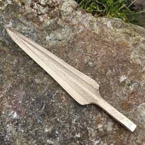 Puede incluir: Una punta de lanza de color bronce con una hoja larga y puntiaguda. La punta de lanza est&aacute; sobre una gran roca texturizada con hierba verde en el fondo. Un artefacto hist&oacute;rico.