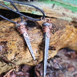 May include: Two black cord necklaces with copper wire wrapped around the top of each pendant. The pendants are made from large, rusty nails.