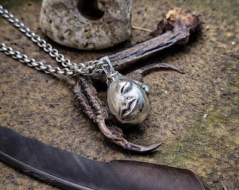 May include: A silver pendant necklace featuring a stylized face, resting on a bird's claw. A dark feather and a stone with a hole are also present. The background is a textured, earthy surface.