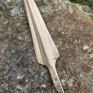 Puede incluir: Una punta de lanza de color bronce con una hoja larga y puntiaguda y un v&aacute;stago corto y rectangular. La punta de lanza est&aacute; sobre una piedra rugosa, gris y marr&oacute;n, con hierba verde al fondo.