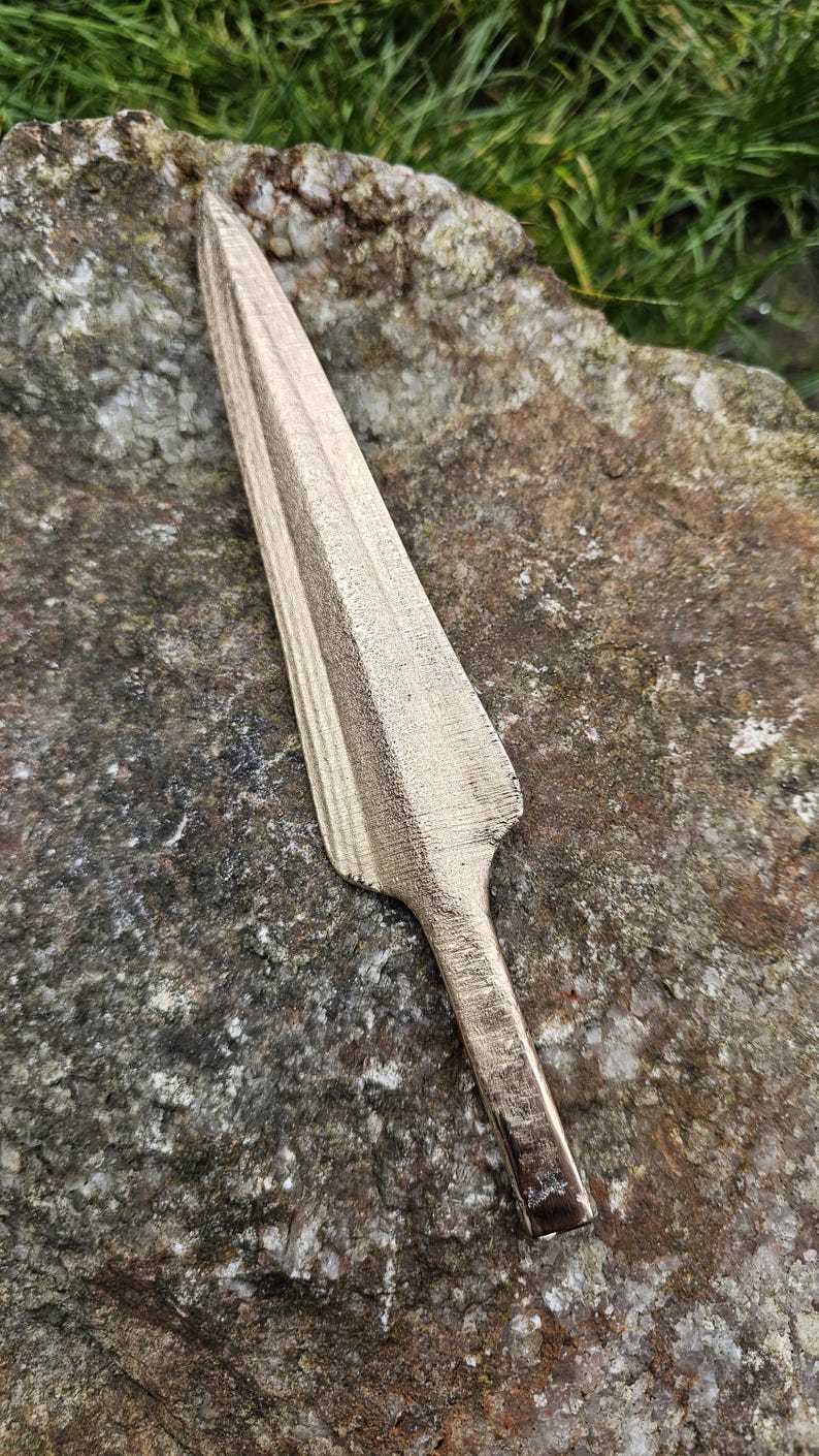 Puede incluir: Una punta de lanza de color bronce con una hoja larga y puntiaguda y un v&aacute;stago corto y rectangular. La punta de lanza est&aacute; sobre una piedra rugosa, gris y marr&oacute;n, con hierba verde al fondo.