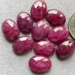 Puede incluir: Un conjunto de gemas de rubí ovaladas y facetadas en tonos rojos y rosas. Las gemas están dispuestas sobre una superficie gris claro, con una moneda para comparar el tamaño. Las gemas tienen una superficie pulida y reflectante.