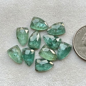 May include: Ten faceted green emerald gemstones, each with a pointed tip, arranged on a white surface. A US quarter is included for scale.