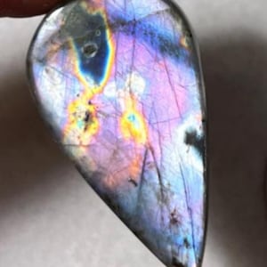 Puede incluir: Una piedra preciosa de labradorita en forma de lágrima pulida. La piedra muestra un vibrante juego de colores, con destellos iridiscentes de púrpura, azul y amarillo. La gema se sostiene entre los dedos, mostrando su belleza natural y patrones únicos.