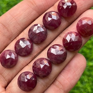 May include: A collection of faceted, round, deep red gemstones. The gems are held in a hand, showcasing their polished surfaces and intricate cuts. The stones have a rich, saturated color, and are likely rubies.