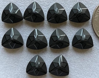 May include: A collection of ten black, triangular-shaped gemstones. Each gem has a faceted design with a central point and geometric patterns. The gems are arranged on a light-colored surface, with a US quarter for size comparison.