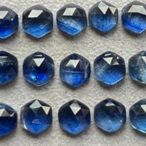 May include: Rows of faceted, hexagonal blue gemstones. The gems vary in shades of blue, from light to deep indigo. The facets catch the light, creating a sparkling effect. The gemstones are arranged on a neutral surface.