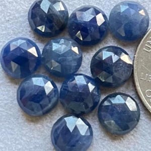 Könnte beinhalten: Zehn facettierte blaue Saphir-Edelsteine, jeder mit einem einzigartigen Schliff und einer einzigartigen Form. Die Edelsteine sind in einer Gruppe auf einer weißen Oberfläche angeordnet.