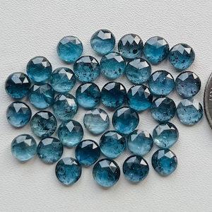 May include: A collection of 30 round, faceted, blue aquamarine gemstones. The stones are approximately 6mm in diameter and have a light blue color with some inclusions. The stones are arranged in a pile on a white surface.