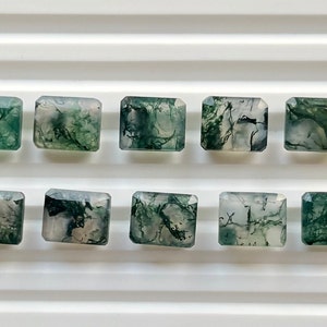 May include: Ten rectangular moss agate gemstones with a polished finish. The stones are translucent with green, white, and black inclusions, arranged in two rows on a white surface. Each stone has a rectangular shape with cut corners.