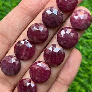May include: A collection of faceted, round, deep red gemstones. The gems are held in a hand, with a green background visible. The gems have a polished, reflective surface, and are likely rubies, suitable for jewelry making.