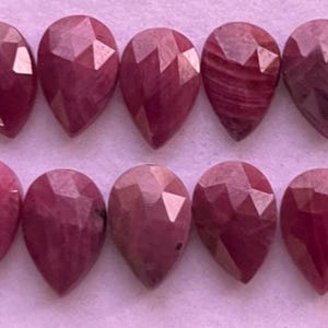 Puede incluir: Diez gemas facetadas en forma de lágrima, en varios tonos de rojo intenso y rosa, dispuestas en dos filas. Las gemas tienen una superficie pulida y reflectante, con patrones internos visibles. El efecto general es de color y textura ricos.