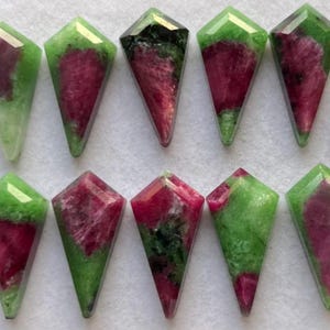 May include: A collection of ten gemstone cabochons. Each gem is a kite shape with a faceted top. The stones feature a mix of deep red and green colors. The gems are arranged on a light-colored surface.