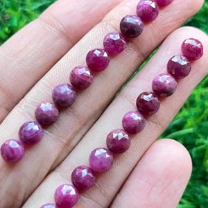 May include: A strand of faceted ruby beads, each bead is a deep pink colour and has a faceted, rounded shape. The beads are arranged in a line on a hand, with the beads on the left side of the image slightly larger than the beads on the right side.