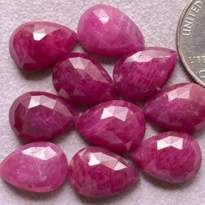 Puede incluir: Una colección de rubíes facetados en forma de lágrima, en varios tonos de rosa y rojo. Las piedras preciosas están dispuestas juntas, con una moneda para comparar el tamaño. Los rubíes tienen una superficie pulida y reflectante.
