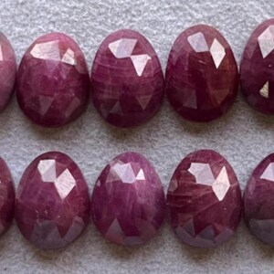 Puede incluir: Se muestran diez piedras preciosas rojas oscuras, facetadas y de forma ovalada. Las gemas tienen una superficie pulida y reflectante y están dispuestas en dos filas. Se incluye una moneda para comparar el tamaño. Las piedras preciosas son probablemente rubíes.