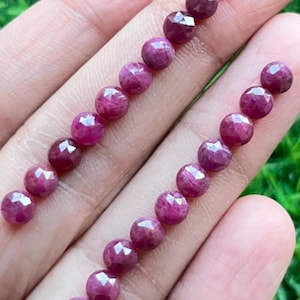 Puede incluir: Una hebra de cuentas de rubí facetadas, cada cuenta es de color rosa intenso y tiene una forma geométrica facetada. Las cuentas están dispuestas en línea sobre una mano, con las cuentas del lado izquierdo de la imagen ligeramente más grandes que las cuentas del lado derecho.