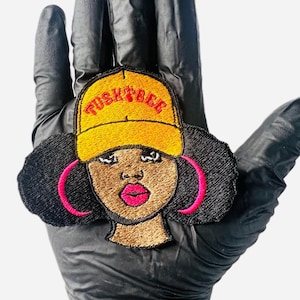 May include: Embroidered patch featuring a cartoon illustration of a person with black hair, wearing a yellow baseball cap with the text "Tusky Bee" in red. The person is wearing pink hoop earrings and has brown skin.