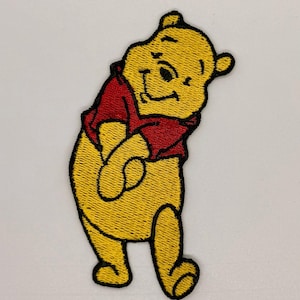 May include: Embroidered patch featuring a smiling yellow bear wearing a red shirt. The bear has black eyes and a black outline. The patch is likely for clothing or accessories.